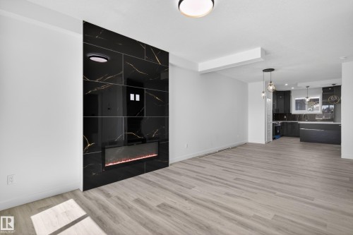 Spacious interior featuring a floor-to-ceiling black tile accent wall with gold veining, an integrated linear electric fireplace, wood-finish flooring, recessed lighting, and a kitchen with dark cabinetry and pendant lighting - 408 Crystal Creek Link, Leduc, AB - Indoor