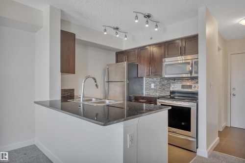 The kitchen features dark wood cabinetry, stainless steel appliances, a grey stone backsplash, and granite countertops - 415 12035 22 Avenue, Edmonton, AB - Indoor Photo Showing Kitchen With Double Sink