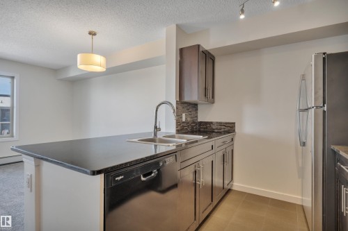 The kitchen features dark cabinetry, a double basin sink, and a dishwasher integrated into the island - 415 12035 22 Avenue, Edmonton, AB - Indoor Photo Showing Kitchen With Double Sink