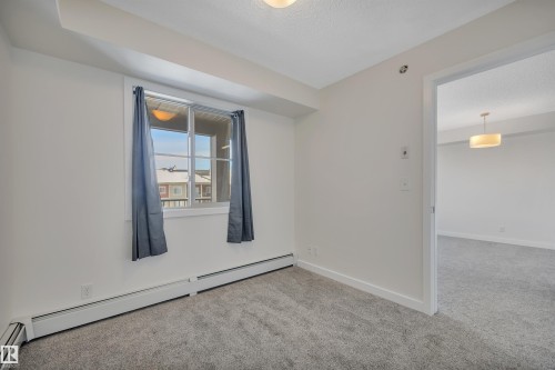 This room features light gray carpet flooring, white walls, and a window with dark gray curtains, offering views of exterior architecture - 415 12035 22 Avenue, Edmonton, AB - Indoor Photo Showing Other Room
