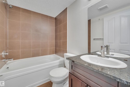 Bathroom featuring a bathtub with a shower, brown tiled walls, a toilet, a vanity with a dark countertop and an undermount sink, and a large mirror - 415 12035 22 Avenue, Edmonton, AB - Indoor Photo Showing Bathroom