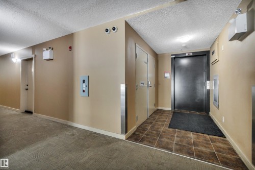 The property features a hallway with carpeted flooring leading to a tiled area, a black elevator door, and light beige walls - 415 12035 22 Avenue, Edmonton, AB - Indoor Photo Showing Other Room