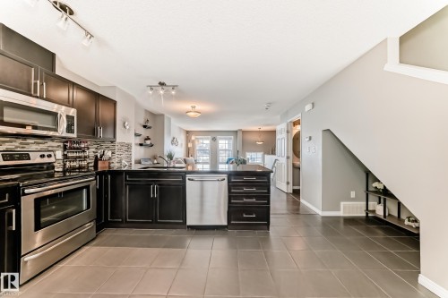 The kitchen features dark wood cabinetry, stainless steel appliances, and a tiled backsplash, with a tiled floor extending throughout the space - 86 4029 Orchards Drive, Edmonton, AB - Indoor Photo Showing Kitchen With Upgraded Kitchen