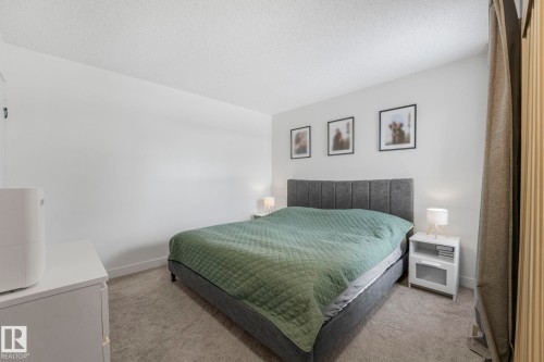 This inviting room features soft gray carpeting, a textured upholstered bed frame, and modern white nightstands with lamps - 33 4403 Riverbend Road, Edmonton, AB - Indoor Photo Showing Bedroom