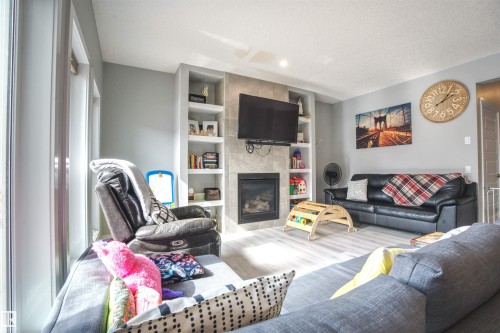 Living area featuring light-colored flooring, a fireplace with a stone surround, built-in shelving, and large windows providing natural light - 20503 99 Avenue, Edmonton, AB - Indoor Photo Showing Living Room With Fireplace
