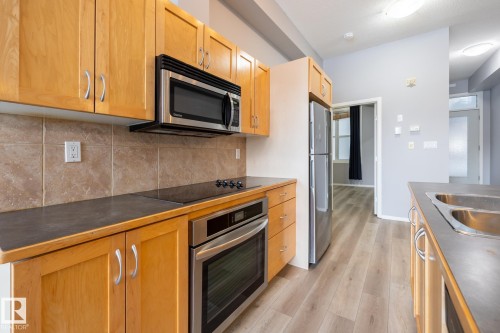 The kitchen features light wood cabinetry, stainless steel appliances, a tiled backsplash, and dark countertops - 210 10147 112 Street, Edmonton, AB - Indoor Photo Showing Kitchen