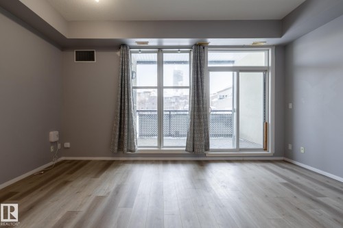 This room features light-colored flooring and a large window with a glass sliding door, providing access to an outdoor area with a black railing - 210 10147 112 Street, Edmonton, AB - Indoor Photo Showing Other Room