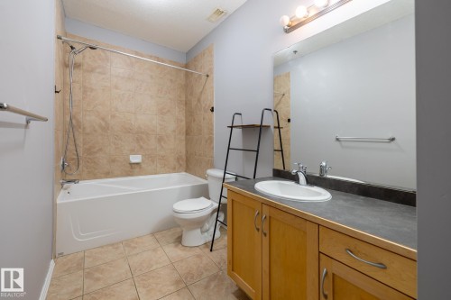 Bathroom featuring a white bathtub with tiled surround, a toilet, a vanity with a light wood cabinet, and a large mirror with overhead lighting - 210 10147 112 Street, Edmonton, AB - Indoor Photo Showing Bathroom