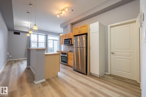 The property features an open-concept living space with light wood-style flooring, a kitchen area with light wood cabinetry and stainless steel appliances including a refrigerator, oven, and microwave - 210 10147 112 Street, Edmonton, AB - Indoor Photo Showing Kitchen With Stainless Steel Kitchen