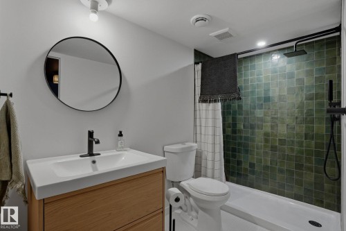 Bathroom featuring a light wood vanity with a white countertop and rectangular sink, a black framed circular mirror, and a shower with green subway tiles and matte black fixtures - 9228 100 Avenue, Edmonton, AB - Indoor Photo Showing Bathroom