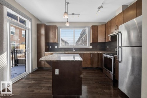 The kitchen features dark wood flooring, brown cabinetry, a center island with a light-colored countertop, and stainless steel appliances - 2105 Casselman Link Link, Edmonton, AB - Indoor Photo Showing Kitchen With Stainless Steel Kitchen With Upgraded Kitchen