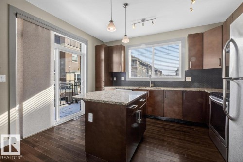 The kitchen features dark wood cabinetry, a granite-topped island, stainless steel appliances, and a tiled backsplash - 2105 Casselman Link Link, Edmonton, AB - Indoor Photo Showing Kitchen With Stainless Steel Kitchen With Upgraded Kitchen