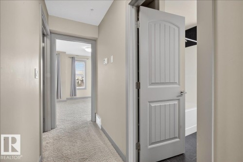 Hallway with light-colored walls and plush carpeting, featuring a view into a carpeted room with a window providing natural light, and a partially open door revealing dark floor tiles - 2105 Casselman Link Link, Edmonton, AB - Indoor Photo Showing Other Room