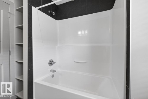 Bathroom with a white tub and shower combination featuring chrome fixtures, set against dark tiled walls and a built-in shelving unit - 2105 Casselman Link Link, Edmonton, AB - Indoor Photo Showing Bathroom
