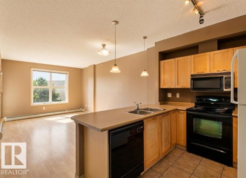 This open concept living space features a kitchen with light wood cabinetry, a double basin sink, and black appliances, including a microwave, oven, and dishwasher - 1-204 4245 139 Avenue, Edmonton, AB - Indoor Photo Showing Kitchen With Double Sink