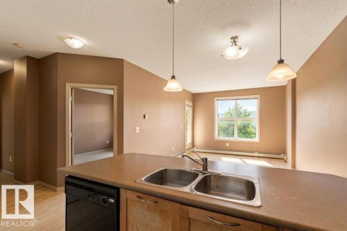The property features a kitchen island with a double basin sink and dishwasher, pendant lighting, and hardwood flooring - 1-204 4245 139 Avenue, Edmonton, AB - Indoor Photo Showing Kitchen With Double Sink