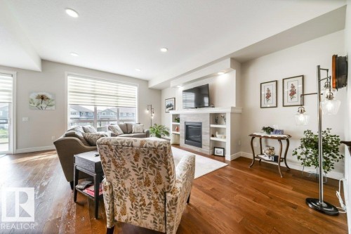 The living room features rich wood flooring, recessed lighting, and large windows with blinds - 3 8050 Orchards Green, Edmonton, AB - Indoor Photo Showing Living Room With Fireplace