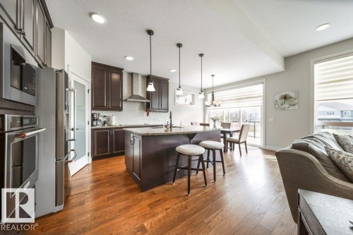 This open-concept kitchen features rich wood flooring, dark cabinetry, and stainless steel appliances - 3 8050 Orchards Green, Edmonton, AB - Indoor Photo Showing Kitchen With Upgraded Kitchen