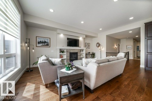 Living room featuring hardwood floors, a fireplace with a stone surround, recessed lighting, and a large window with horizontal blinds - 3 8050 Orchards Green, Edmonton, AB - Indoor Photo Showing Living Room With Fireplace