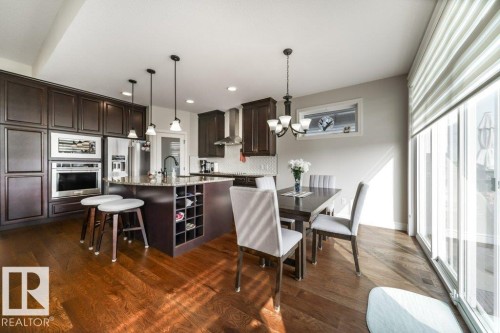 Kitchen and dining area featuring hardwood floors, dark wood cabinetry, a kitchen island with seating, stainless steel appliances, and large sliding glass doors - 3 8050 Orchards Green, Edmonton, AB - Indoor