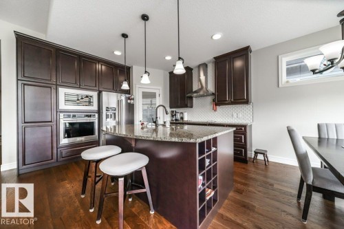 The kitchen features dark wood cabinetry, stainless steel appliances, a granite countertop island with integrated shelving, and hardwood flooring - 3 8050 Orchards Green, Edmonton, AB - Indoor Photo Showing Kitchen With Upgraded Kitchen