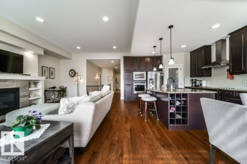 This open concept living space features hardwood floors, a kitchen with dark wood cabinetry, stainless steel appliances, and a central island with pendant lighting - 3 8050 Orchards Green, Edmonton, AB - Indoor With Fireplace