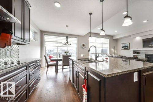 The kitchen features dark wood cabinetry, a large kitchen island with granite countertops, and hardwood flooring - 3 8050 Orchards Green, Edmonton, AB - Indoor Photo Showing Kitchen With Double Sink With Upgraded Kitchen