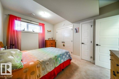 This interior space features light-colored carpeting and a window with blinds, framed by sheer, colorful curtains - 3 8050 Orchards Green, Edmonton, AB - Indoor Photo Showing Bedroom