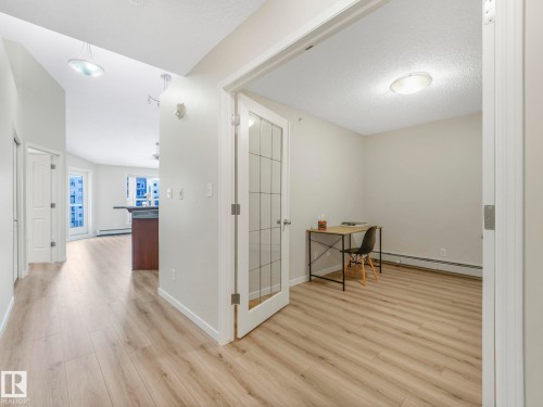 The property offers light-toned flooring throughout, a room with a clear glass paneled door, and a hallway leading to a bright living area - 515 10311 111 Street, Edmonton, AB - Indoor Photo Showing Other Room