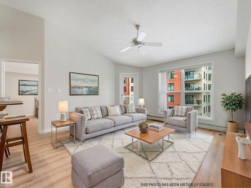 The living area features light-toned flooring, a ceiling fan, and a large window providing views of nearby buildings. This photo includes virtual staging. - 515 10311 111 Street, Edmonton, AB - Indoor Photo Showing Living Room