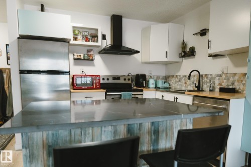 The kitchen features a large kitchen island with a dark countertop and a paneled base, stainless steel appliances, white cabinetry, and a geometric pattern backsplash - 303 10432 76 Avenue, Edmonton, AB - Indoor Photo Showing Kitchen With Upgraded Kitchen