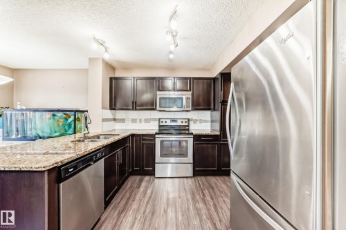 The kitchen features dark wood cabinetry, granite countertops, and stainless steel appliances, including a refrigerator, stove, and microwave - 23 445 Brintnell Boulevard Nw, Edmonton, AB - Indoor Photo Showing Kitchen With Double Sink With Upgraded Kitchen