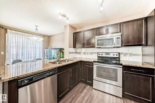 The kitchen features dark wood cabinetry, stainless steel appliances, granite countertops, and a tile backsplash - 23 445 Brintnell Boulevard Nw, Edmonton, AB - Indoor Photo Showing Kitchen With Double Sink With Upgraded Kitchen
