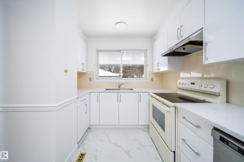 The kitchen features white cabinetry, light-colored countertops, and a window with blinds overlooking a snowy outdoor scene - 6411 149 Avenue Nw, Edmonton, AB - Indoor Photo Showing Kitchen