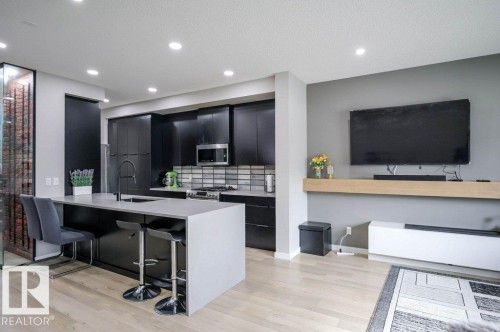 The open concept living area features a kitchen with dark cabinetry, a light-colored island with seating, a stainless steel microwave, and a tiled backsplash - 266 Charlesworth Drive, Edmonton, AB - Indoor