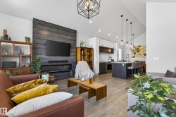 Open concept living area featuring light-toned flooring, a modern fireplace with a dark wood-paneled wall, and a kitchen with white cabinetry and an island - 