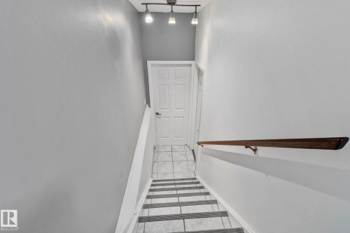 Staircase featuring a wooden handrail, track lighting, and a white paneled door at the bottom - 17082 67 Avenue Nw, Edmonton, AB - Indoor Photo Showing Other Room