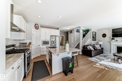 The open concept living area features light hardwood flooring, a kitchen with white cabinetry and stainless steel appliances, and a living room with a white fireplace surround - 