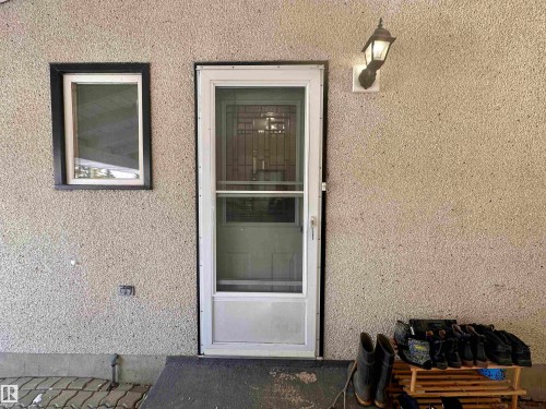 The property features a textured exterior wall, a white framed storm door with a decorative glass panel, and a wall-mounted exterior light fixture - 9915 95 Street, Westlock, AB - Outdoor With Exterior