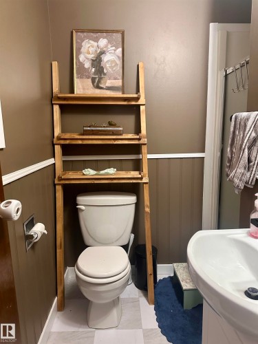 The bathroom features a white toilet, a white sink, and a wooden over-the-toilet storage unit with three shelves - 9915 95 Street, Westlock, AB - Indoor Photo Showing Bathroom