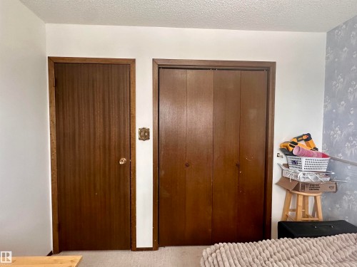 This room features a closet with bi-fold doors and a solid wood door, complemented by a textured ceiling and a wall with floral wallpaper - 9915 95 Street, Westlock, AB - Indoor Photo Showing Bedroom