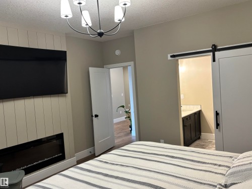 This room features a modern chandelier, a wall-mounted television above an electric fireplace, and a barn door entry to an additional space - 137 Graybriar Drive, Stony Plain, AB - Indoor Photo Showing Bedroom