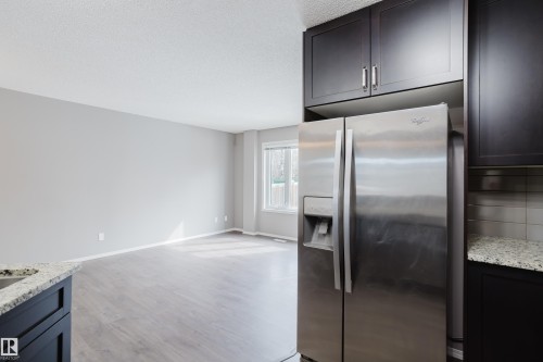 The kitchen features dark cabinetry, granite countertops, and a stainless steel refrigerator, overlooking a living area with light-colored walls and wood-look flooring - 14007 164 Avenue, Edmonton, AB - Indoor Photo Showing Kitchen