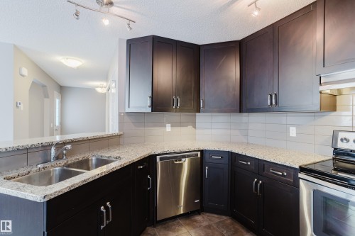 The kitchen features dark wood cabinetry, light speckled countertops, a double stainless steel sink, and stainless steel appliances, including a dishwasher and oven with a stovetop - 14009 164 Avenue, Edmonton, AB - Indoor Photo Showing Kitchen With Double Sink With Upgraded Kitchen