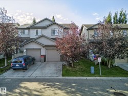 The property features light gray siding, two garage doors, and a well-maintained lawn with mature trees - 