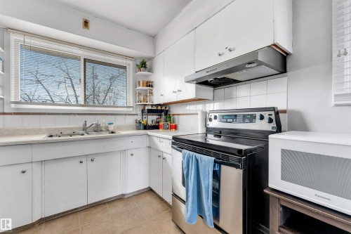 The kitchen features a window with blinds, white cabinetry, a stainless steel electric stove, a range hood, and a microwave - 12908 126 Street, Edmonton, AB - Indoor Photo Showing Kitchen