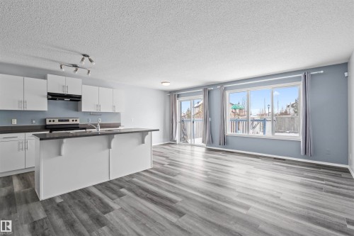 This open concept living area features light grey flooring, a kitchen with white cabinetry and dark countertops, and a sliding glass door leading to an outdoor space - 11542 18A Avenue, Edmonton, AB - Indoor Photo Showing Kitchen With Double Sink