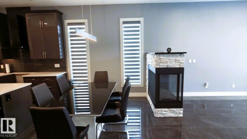 This inviting interior features a contemporary corner fireplace with a stone surround, dark tile flooring, and a modern kitchen with dark cabinetry - 1781 Melrose Crescent, Edmonton, AB - Indoor Photo Showing Dining Room With Fireplace