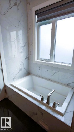 Bathroom featuring a built-in bathtub with a brushed nickel faucet, a window with a blind, and white marble-patterned wall tiles - 1781 Melrose Crescent, Edmonton, AB - Indoor Photo Showing Bathroom