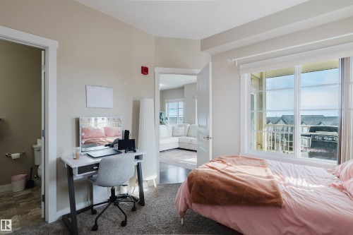 The room features a window with a view of exterior rooftops, a doorway leading to a bathroom with a toilet and tiled flooring, and a doorway opening to a living area with a white sofa - 402 3670 139 Avenue, Edmonton, AB - Indoor Photo Showing Bedroom